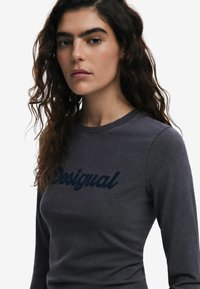 Dlhý rukáv sivé tričko s okrúhlym výstrihom a výrazným tmavomodrým logom "Desigual" na prednej strane.