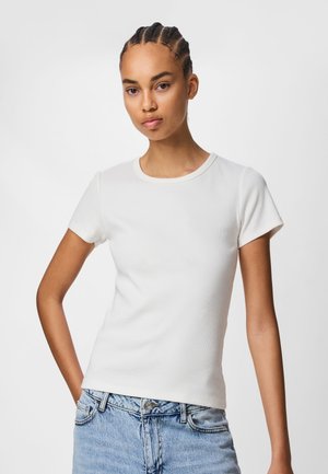 Mladá žena s cornrow vrkočmi, oblečená v priliehavé biele krátke tričko a svetlomodré džínsy, stojaca na jednoduchom pozadí.