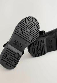 Pantofi negri din cauciuc, cu o talpă texturată cu modele în formă de stea și un design cu bretele. Partea inferioară prezintă numele brandului "NEXT."