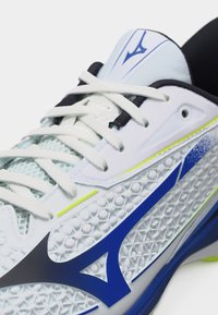 Hvid sportssko med blå og sorte geometriske accenter, tekstureret overdel, hvide snørebånd og en lille gul stribe nær mellemsålen.