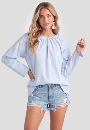 Frau mit welligem blonden Haar trägt eine hellblaue langärmelige Bluse, ausgewaschene Denimshorts und Quastenohrringe vor neutralem Hintergrund.