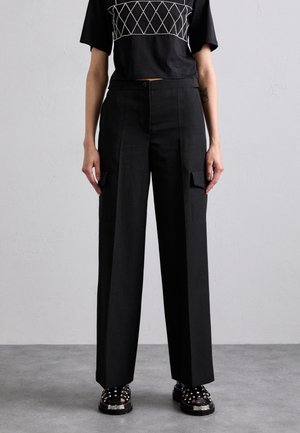 Osoba nosząca czarne spodnie o wysokim stanie i szerokich nogawkach z klapkowymi kieszeniami, zestawione z czarnymi platformami z ćwiekami i krótkim czarnym topem.