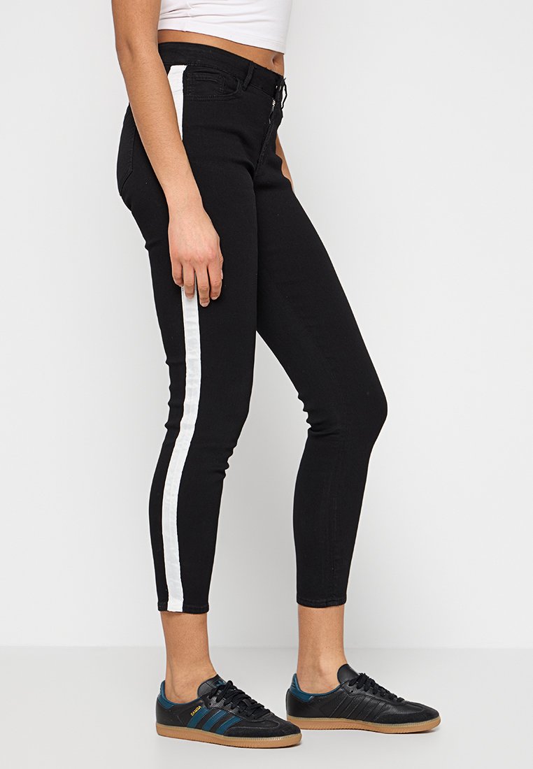 Jeans en denim noir avec une jambe fuselée, dotés d'une bande latérale blanche. La taille a un bouton de fermeture et ils offrent une coupe taille intermédiaire.