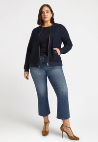 Jachetă de bombardier navy cu țesătură texturată și închidere cu fermoar, asortată cu un top navy și blugi scurți de culoare albastru deschis. Tocuri din piele lacuită maro.