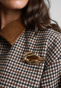 Spilla d'oro a forma di foglia su un cappotto houndstooth con colletto in velluto a coste, caratterizzato da motivi crema, marrone e verde.