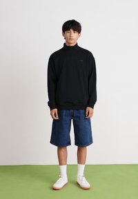 Sudadera negra con cremallera, pantalones cortos de mezclilla azules, calcetines blancos hasta la mitad de la pantorrilla y zapatillas blancas con suelas marrón claro. Tejido suave y corte relajado.