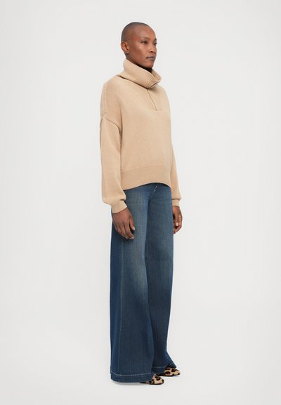 Beige Strickpullover mit hohem Kragen, kombiniert mit weiten blauen Jeans. Das Outfit besticht durch eine lockere Passform und strukturierte Strickdetails.