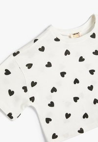 Hvid kortærmet T-shirt med et gentaget mønster af små sorte hjerter. Lavet af blødt stof med en rund halsudskæring.