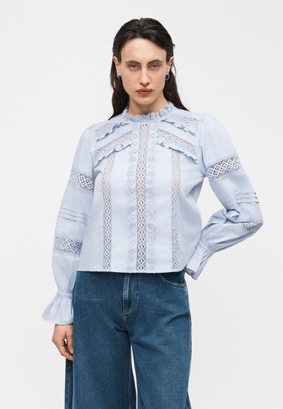 Νεαρή γυναίκα με σκουρόχρωμα μαλλιά φοράει ανοιχτό μπλε μπλούζα με δαντελένιες πιέτες, βολάν και μακριά φουσκωτά μανίκια, σε συνδυασμό με μπλε τζιν.