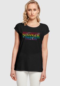 Svart T-skjorte med korte oppbrettede ermer, med en fargerik "STRANGER THINGS" tekstgrafikk i en retro, pikselert stil.