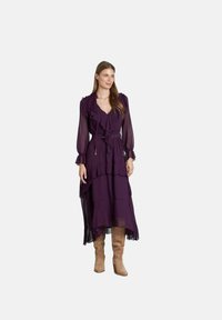 Vestido maxi morado de volantes y con varias capas en tejido ligero y fluido; presenta mangas largas con volantes y una cintura ceñida con un cinturón. Botas marrones hasta la rodilla.