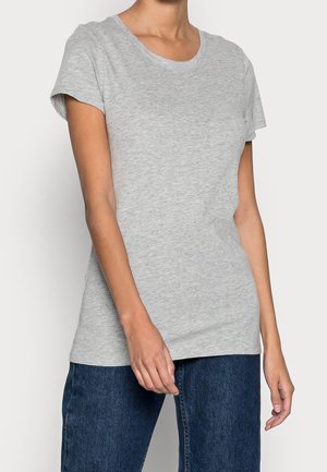 Person iført en ensfarvet lysegrå kortærmet t-shirt og mørkeblå jeans, stående mod en ensfarvet lys baggrund.
