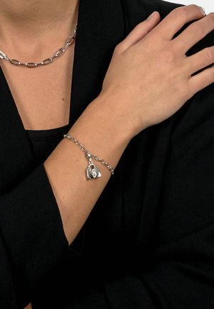 Silberarmband mit einem Kettendesign und einem herzförmigen Anhänger sowie einem klaren Steinakzent, der an einem nackten Handgelenk vor einem dunklen Hintergrund getragen wird.