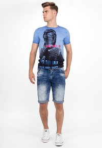 Blauwe graphic t-shirt met een ontwerp van een gevlochten kapsel en tekst, gecombineerd met lichte denim shorts en witte sneakers.