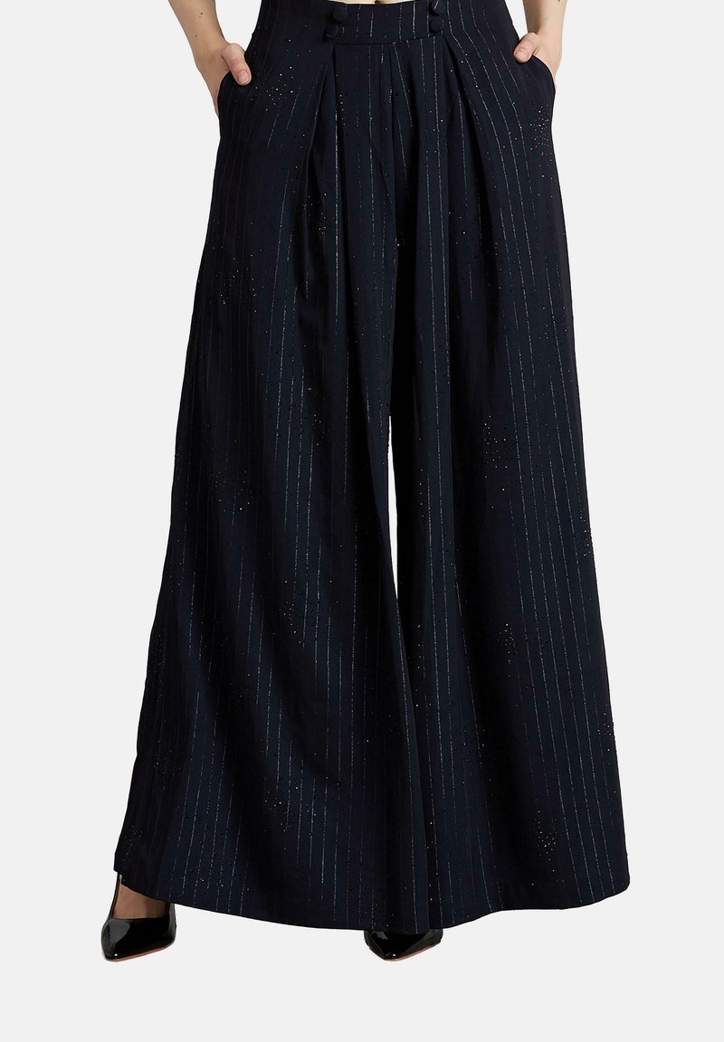 Vrouw in hoogwater-wijdebroek in marineblauw met verticale streepjes en handen in de zakken, gecombineerd met zwarte puntige hakken.