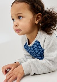 Chemise grise à manches longues avec un panneau sur la poitrine à motif bleu ; cheveux bouclés ; expression concentrée ; mains posées sur une surface.