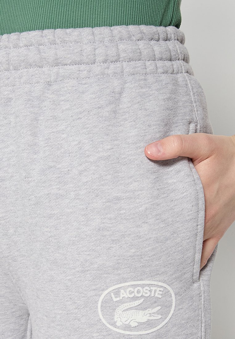 Szare bawełniane dresy z elastycznym paskiem w talii i boczną kieszenią, z białym haftowanym logo Lacoste na lewej nodze.