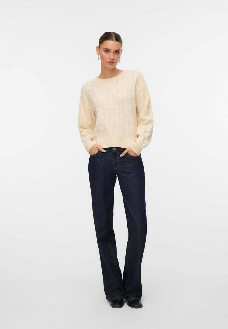 Beige gestrickter Pullover mit strukturiertem Muster und Rundhalsausschnitt, kombiniert mit dunkelblauen Jeans und schwarzem Schuhwerk. Lockerer Sitz, lange Ärmel.