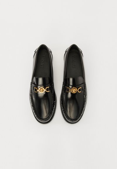 Svarta läderloafers med en accent av guldkedja och rund medaljongdetalj. Slät textur, rund tå och sömnad runt sulan.