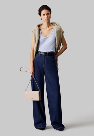 Femme portant un gilet en tricot beige sans manches sur un débardeur bleu clair, jean large et foncé, ceinture noire, tenant un sac à main rose clair.