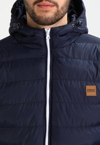 Marineblauwe pufferjack met een witte rits, verstelbare capuchon en gestikte paneelontwerpen. Inclusief een bruin leren label aan de voorkant.