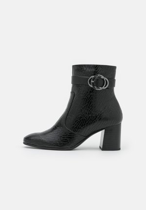 Bottine noire avec motif en cuir texturé, talon bloc, bout pointu et une bride avec une boucle double anneau autour de la cheville.