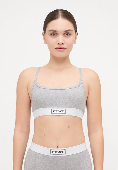 Harmaa puuvillainen bralette ohuiden hihnojen ja valkoisen elastisen nauhan kanssa, jossa on musta logotagi. Yksinkertainen, istuva muotoilu, jossa on sileä rakenne.