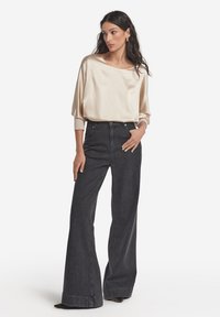 Beżowa satynowa bluzka z szerokim, łodziowym dekoltem i długimi rękawami; w połączeniu z wysokim stanem ciemnych, dzwonowatych dżinsów; gładka faktura i luźny krój.