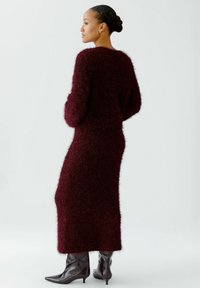 Burgunderfarbenes, flauschiges Strickkleid mit langen Ärmeln und figurbetonter Silhouette. Paar schwarze High-Heels-Stiefel. Strukturierte, gemütliche Optik.