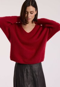 Czerwony sweter oversize z dekoltem w serek, uszyty z miękkiego, puszystego materiału, z szerokimi rękawami, zestawiony z czarną skórzaną spódnicą o wyraźnej fakturze.