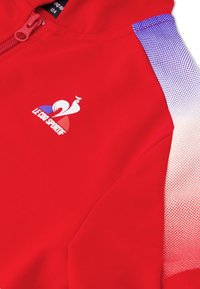 Chaqueta roja con cremallera y logo en el pecho, con detalles de malla degradada en púrpura y blanco en el hombro. Textura de tela suave.