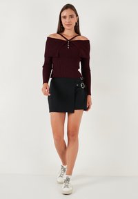 Sieviete, tērpusies bordo krāsas off-shoulder ribota džemperī ar pogām un melnā mini svārkā ar sānu sprādzi, iet uz priekšu baltās kedās.