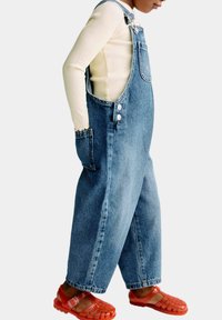 Blauwe denim overall met brede pijpen, voorzien van zijknopen en een voorzak. Gecombineerd met rode plastic sandalen. Lange mouw beige top eronder.