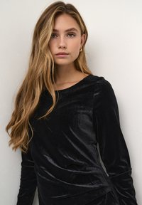 Svart sammetstopp med långa ärmar, med rund halsringning och draperad design. Den släta texturen ger dimension till plagget.