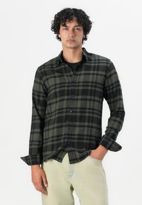Camisa de franela a cuadros verde y negro, de manga larga, diseño de botones, con un bolsillo en el pecho, combinada con denim de colores claros.