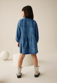 Denimkleid mit blauem Schleifenmuster, geraffter Taille, langen Ärmeln und einem ausgestellten Rock. Kombiniert mit metallischen Stiefeletten und einer glatten Oberfläche.