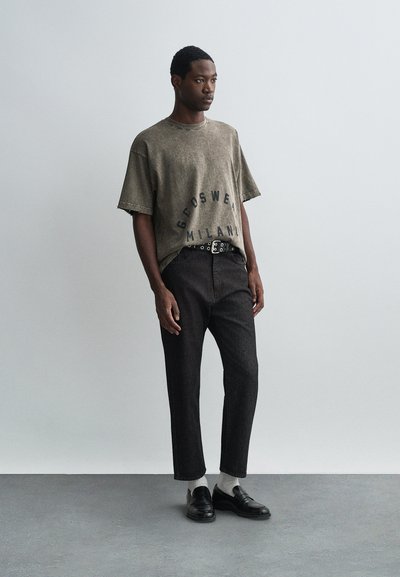 Tricou oversized de culoare verde măsliniu cu imprimeu "Groswear Milan", asortat cu pantaloni din denim închis și mocasini negri, purtând șosete albe.
