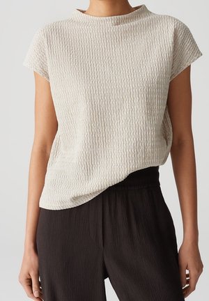 Personne portant un haut beige clair texturé à manches courtes, rentré dans un pantalon noir ample taille haute, debout devant un fond uni clair.