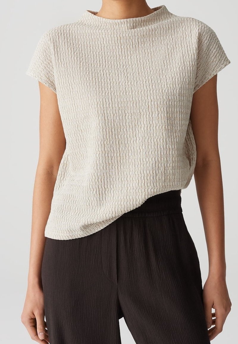Personne portant un haut beige clair texturé à manches courtes, rentré dans un pantalon noir ample taille haute, debout devant un fond uni clair.