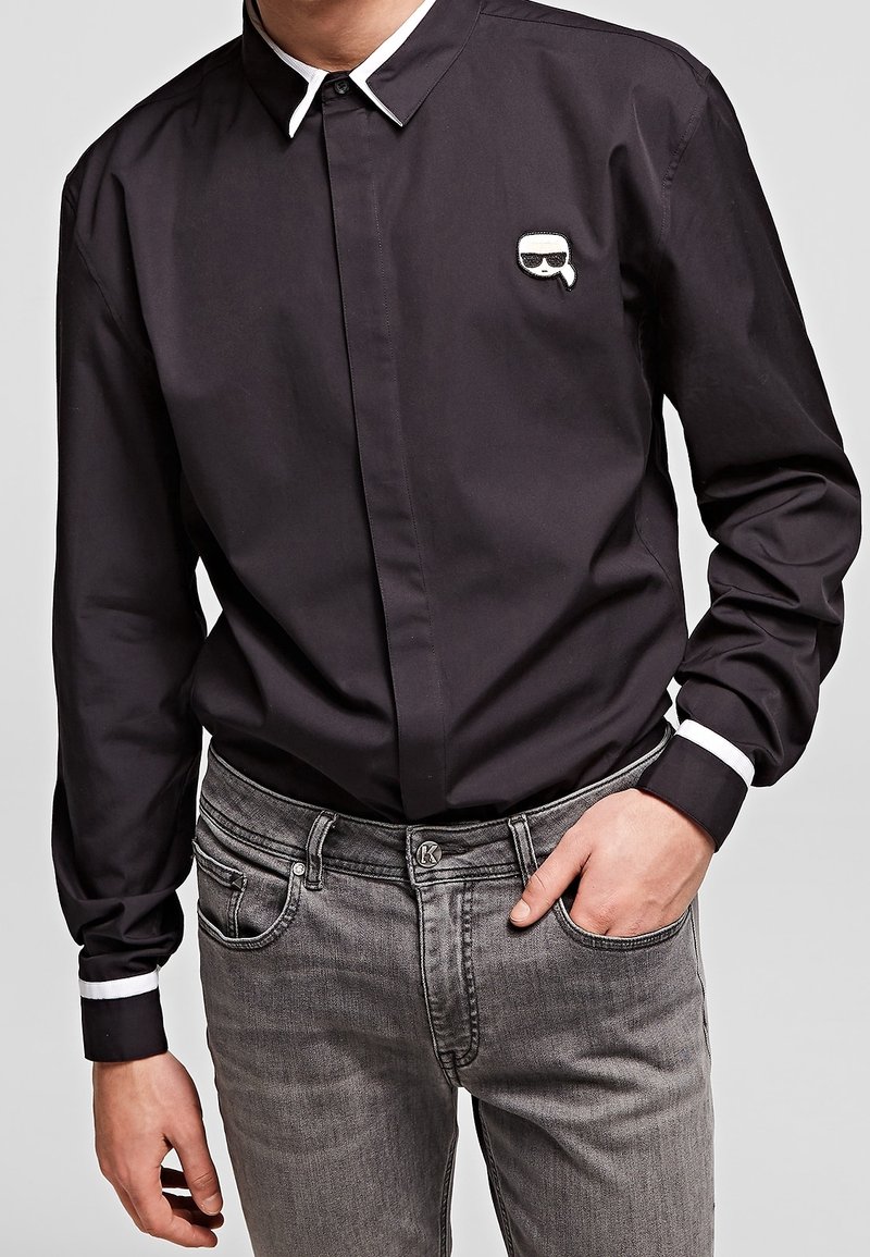 Černá košile s dlouhým rukávem a bílým límcem a manžetami, s malým grafickým logem na hrudi. Tkanina vypadá hladce a těsně.