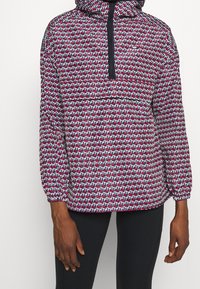 Pullover-Jacke mit Kapuze, mit einem geometrischen Muster in Rot, Weiß und Blau. Elastische Bündchen und eine halbe Reißverschlussöffnung. Leichtes Material.