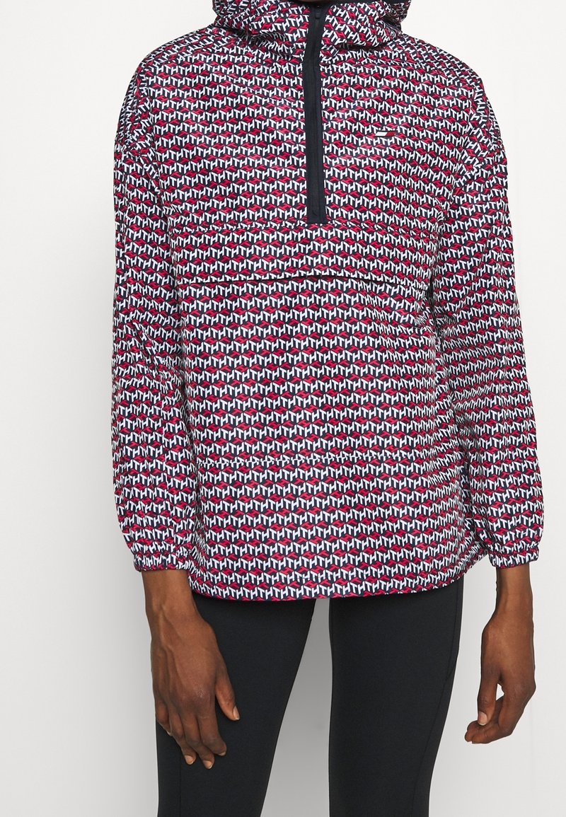 Pullover-Jacke mit Kapuze, mit einem geometrischen Muster in Rot, Weiß und Blau. Elastische Bündchen und eine halbe Reißverschlussöffnung. Leichtes Material.