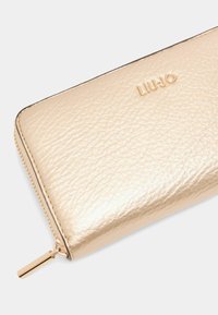 Portafoglio in pelle con texture dorata e chiusura a zip, dotato di una leggera embossatura del logo e di un tirazip metallico.