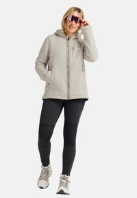 Veste en polar gris clair avec fermeture éclair à l'avant, poches latérales et capuche, associée à des leggings noirs et des chaussures de randonnée de couleur claire.