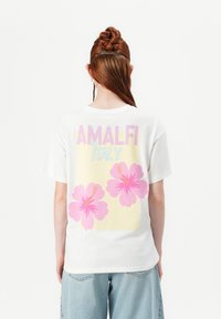 Balts kokvilnas t-krekls ar uzrakstu "AMALFI Itālija" un diviem rozā hibiska ziediem uz dzeltena fona. Brīva piegriezuma, īsām piedurknēm.