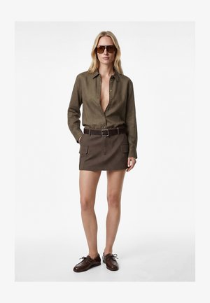 Vrouw draagt een donkerolijfgroen overhemd met knopen, een bruine rok met een riem en voorzakken, donkerbruine veterschoenen en grote zonnebril, staat rechtop.