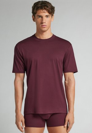 Jonge man met een effen kastanjebruin T-shirt met korte mouwen en bijpassende aansluitende korte broek, staand tegen een neutrale achtergrond.