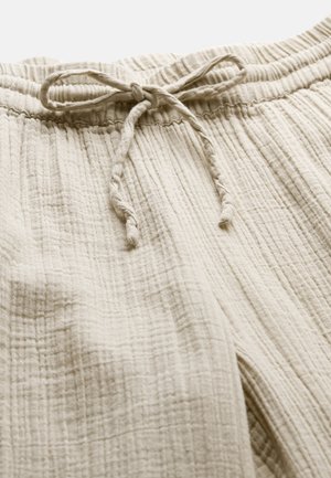 Lysebeige bukser i struktureret stof med elastisk taljebånd og en knyttet snøre foran i midten.