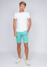 T-shirt blanche à manches courtes associée à un short turquoise retroussé et des baskets blanches, se tenant sur un fond gris clair.