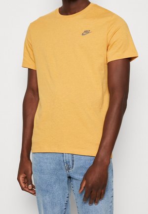 Gelbes Baumwoll-T-Shirt mit kurzen Ärmeln und Rundhalsausschnitt, mit einem kleinen blauen Nike-Logo auf der linken Brust. Kombiniert mit hellen Jeans.